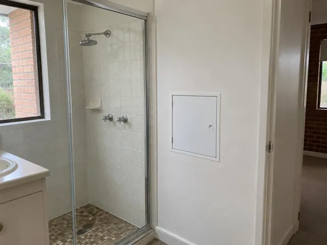 Bathroom wall with a small access panel beside a glass shower in a tiled home interior in Doonside